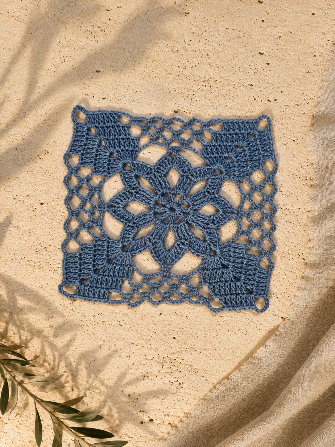 Blue handmade crochet square motif study with a floral centre, openwork sections and defined corner structure on a warm neutral surface.
