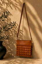 Hanging view of the cognac brown crossbody bag showing the full leather strap, zip-top panel and five rows of leather sequins.