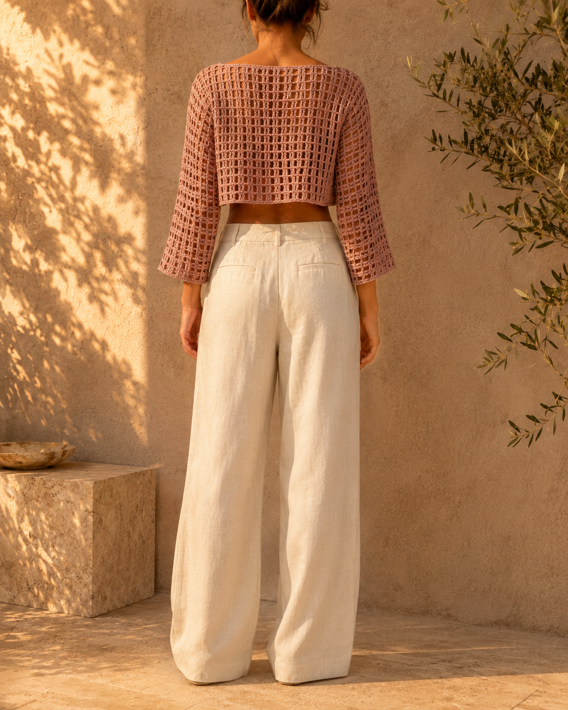 Back view of the long-sleeve openwork crochet crop top styled with high-waisted white trousers.