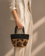 Handmade black and tan leopard-pattern paper yarn handbag with rigid glossy handles, carried by hand with a neutral outfit.