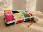 Handmade multicolour crochet blanket folded horizontally on a travertine table beside yarn and a crochet hook.