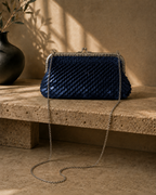 Handmade navy sequin clutch bag carried by hand with a neutral tailored outfit, showing its compact shape and kiss-lock closure.