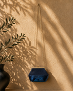 Navy handmade crochet evening bag with partial sequin detailing placed on a warm studio wall with olive-branch shadows.