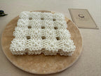 Small square white crochet doily displayed on a neutral stone surface, showing its compact four-by-four motif construction.
