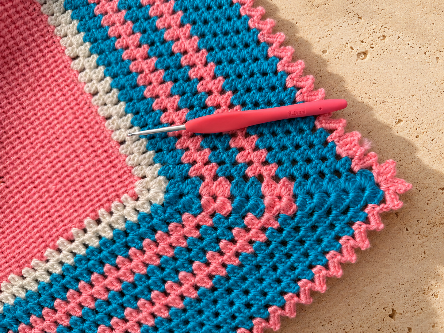 Close-up of the multicolour striped crochet border with a crochet hook resting on the handmade blanket.