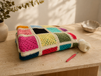 Handmade pink and turquoise Tunisian crochet blanket folded on a travertine table beside yarn and a crochet hook.