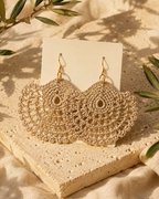 Matching pair of warm sand-beige round crochet earrings with gold-tone hooks displayed on a neutral stone surface.