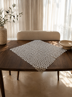 Large white crochet doily displayed diagonally on a dining table, showing its square shape and decorative lace surface.
